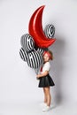 Little child in t-shirt, red headband, black skirt and sneakers. Holding balloons, posing sideways isolated on white. Full length Royalty Free Stock Photo