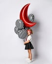 Little child in t-shirt, red headband, black skirt and sneakers. Holding balloons, posing sideways isolated on white. Full length Royalty Free Stock Photo