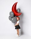 Little child in t-shirt, red headband, black skirt and sneakers. Holding balloons, posing sideways isolated on white. Full length Royalty Free Stock Photo