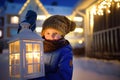Little child is waiting Santa Claus on snowy street of small town on Christmas eve. Boy is holding large lantern with burning Royalty Free Stock Photo