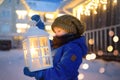 Little Child Is Waiting Santa Claus On Street Of Small Town During Snowfall On Christmas Eve. Boy Is Holding Large Lantern With