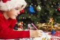 Little child is writing letter to Santa Claus at table. A kid in Santa hat is writing wishlist near the Christmas tree Royalty Free Stock Photo