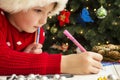 Little child is writing letter to Santa Claus at table. A kid in Santa hat is writing wishlist near the Christmas tree Royalty Free Stock Photo