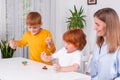 Two little children with a nanny or mother or teacher sit at the table in the room and play with toys