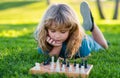 Little clever boy thinking about chess. Kid playing chess, laying on grass in summer park.