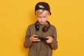 Little concentrated guy wearing casual hirt and cap, playing online video games using mobile phone, posing with headphones around