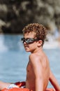 Little curly charismatic boy in swimming goggles sits by the pool and smiles