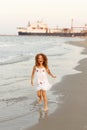 Little curly hair girl jumping on the beach with a white dress, seascape and massive ship background