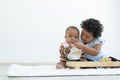 Little cute African older sister try to feeding her newborn sister food while baby playing toys sitting on floor at home.