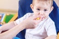 Little cute baby eating on a chair in the kitchen. Mom feeds holding in hand a spoon of porridge