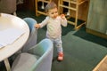 A little cute baby girl is standing with a pencil in his mouth at the children's small low table and child seats in the