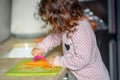 Little cute baby toddler girl in the kitchen peeling carrots with carrot peeler on chopping board. Child help at home