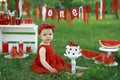 A little cute beautiful girl in a red dress is sitting on the lawn in the park with a festive birthday decor. Royalty Free Stock Photo