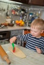 Little cute blond boy in kitchen is learning to cut flour dough with children`s shaped baking knife. Concept of family leasure