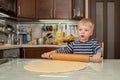 Little cute blond boy in kitchen prepares pie dough by rolling it out on table with rolling pin Royalty Free Stock Photo