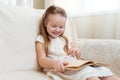 Little cute blond girl reading book siting on a sofa. Child reading, dreaming and imagination development