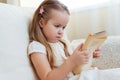 Little cute blond girl reading book siting on a sofa. Child reading, dreaming and imagination development