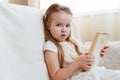 Little cute blond girl reading book siting on a sofa. Child reading, dreaming and imagination development