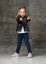 Little cute blond girl in stylish rock style black leather jacket, jeans and sneakers standing and pointing at camera with fingers Royalty Free Stock Photo