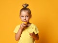 Little cute blond girl in yellow t-shirt holding and licking half of fresh ripe lemon fruit over yellow background