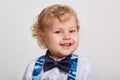 Little cute blonde boy wearing bow tie and blue camouflage suspenders, looking directly at camera with toothy smile, posing Royalty Free Stock Photo