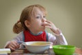 The Little cute blonde curly hair Caucasian girl is seating on the table and eating. She is looking at the window.