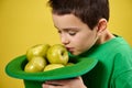 Little cute boy enjoys the scent of green apples in a green Irish leprechaun cap. Face portrait on yellow background. Saint
