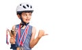 Little cute boy kid wearing bike helmet and winner medals holding winner trophy pointing thumb up to the side smiling happy with