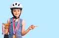 Little cute boy kid wearing bike helmet and winner medals holding winner trophy smiling happy pointing with hand and finger to the