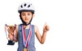 Little cute boy kid wearing bike helmet and winner medals holding winner trophy smiling happy and positive, thumb up doing