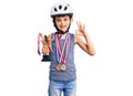 Little cute boy kid wearing bike helmet and winner medals holding winner trophy doing ok sign with fingers, smiling friendly