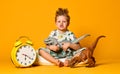 Little cute boy in pajamas holding a toy dinosaur in his hands, sitting on a pillow with an alarm clock. Isolated on a yellow Royalty Free Stock Photo