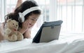 Little cute Caucasian Asian kid smiling lying down on bed with headphone and bear doll using tablet computer to learn online class Royalty Free Stock Photo