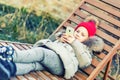 Little cute caucasian blonde toddler girl in red knitted hat and warm jacket lying over lounge and holding smartphon in hand. Ou