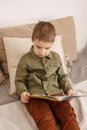 Little and cute caucasian boy reading a book on the bed at home. Interior and clothes in natural earth colors. Cozy Royalty Free Stock Photo