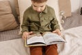 Little and cute caucasian boy reading a book on the bed at home. Interior and clothes in natural earth colors. Cozy Royalty Free Stock Photo