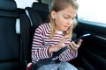 Little cute caucasian girl is driving in car. Kid writing sms, chatting, playing video games in mobile phone. Traveling on road in Royalty Free Stock Photo