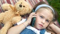 Little cute child girl laying on a blanket on green lawn in summer outdoors with her teddy bear toy talking on mobile phone Royalty Free Stock Photo