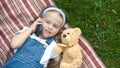 Little cute child girl laying on a blanket on green lawn in summer outdoors with her teddy bear toy talking on mobile phone Royalty Free Stock Photo