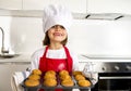 Little and cute girl alone in cook hat and apron presenting and showing tray with muffins smiling happy