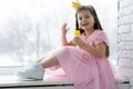 Little cute girl in beautiful dress is sitting near the window at home and blowing soap bubbles. Royalty Free Stock Photo