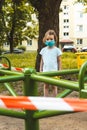 Little cute girl in face mask at closed children`s playground. COVID-19 security and protection measures in the city.