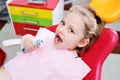 Little cute girl without front milk teeth in red dental chair with electric automatic toothbrush in hands.