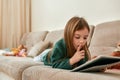 A little cute girl lying on her stomach on a sofa holding a tablet putting a point finger to her opened mouth