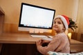 Little cute girl in santa hat sits at table, looks at monitor screen and writes or draws on paper sticker Royalty Free Stock Photo