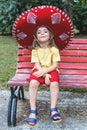 little cute girl wearing a big sombrero is sitting on a red bench in a public park Royalty Free Stock Photo