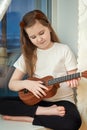 Little cute girl in white t-shirt and black leggings sitting inside of home and playing ukulele Royalty Free Stock Photo