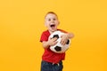 Little cute kid baby boy 3-4 years old, football fan in red t-shirt holding in hand soccer ball isolated on yellow Royalty Free Stock Photo