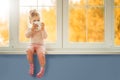 Little cute Kid Girl sitting by window holding cup of hot drink cocoa enjoying autumn forest background. Season Beauty fashion