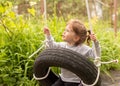 Little cute red-haired girl riding a black wheel swing tied with ropes looks up at